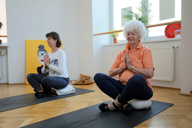 Zwei Seniorinnen sitzen im Schneidersitz auf Yoga-Polstern